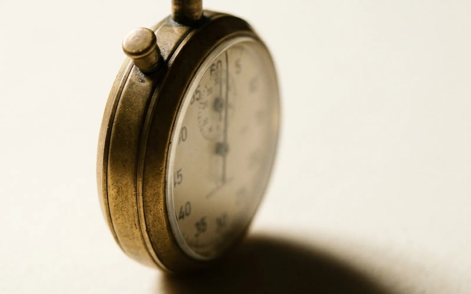 An aged brass mechanical stopwatch resting on cream paper, its dial angled away from the camera.