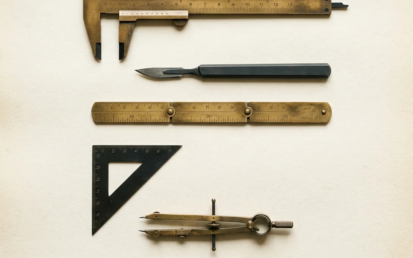 Un pied à coulisse en laiton, un scalpel, un mètre pliant, une équerre et un compas disposés en grille sur du papier crème.